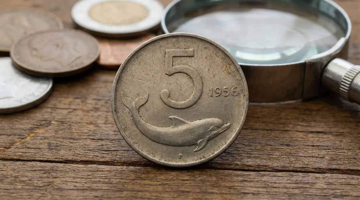 Moneta italiana da 5 lire con delfino e anno 1956, appoggiata su tavolo di legno accanto a una lente d’ingrandimento